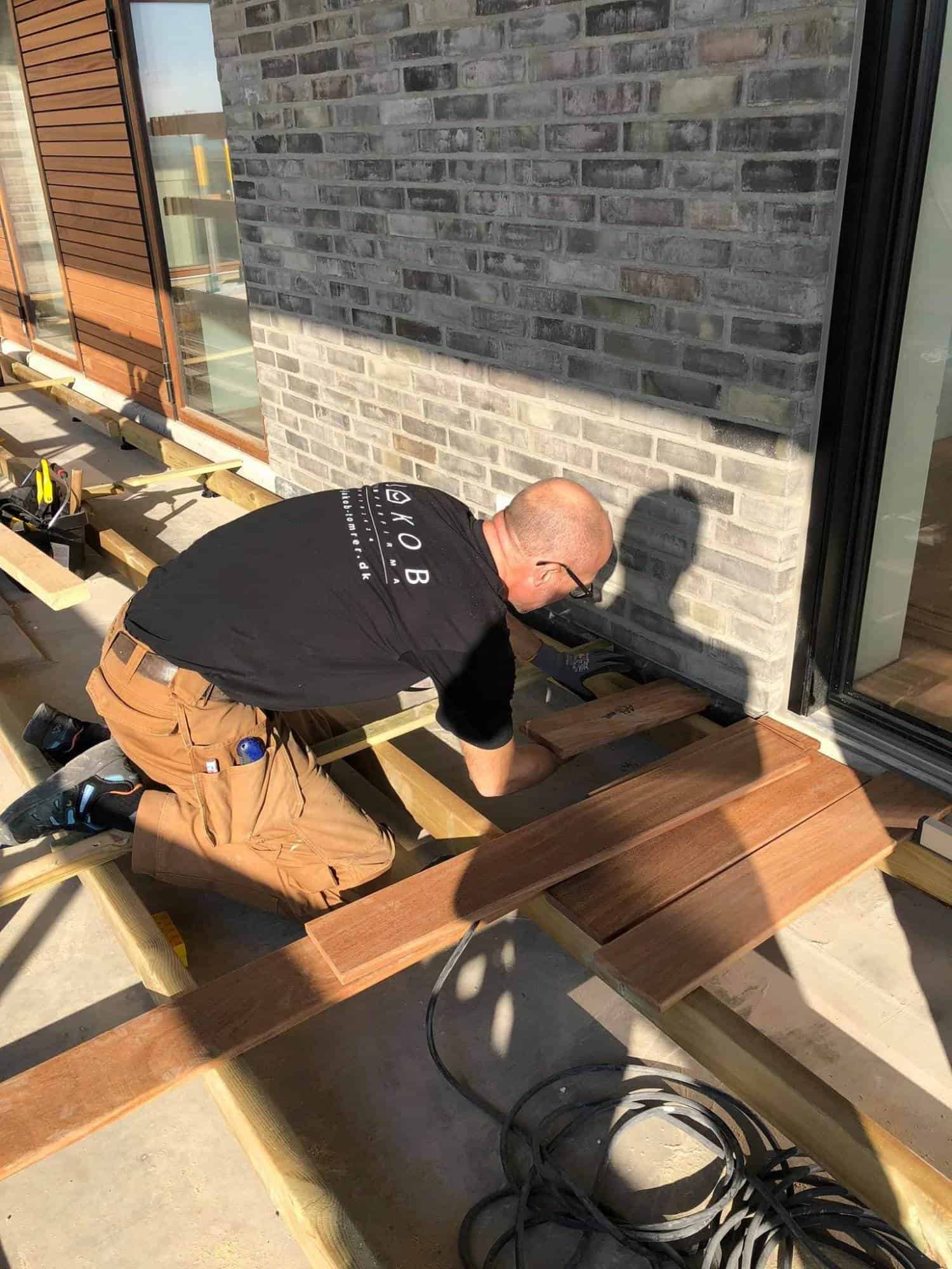 Tømrer arbejder på udendørs terrasse med træplanker og værktøj.