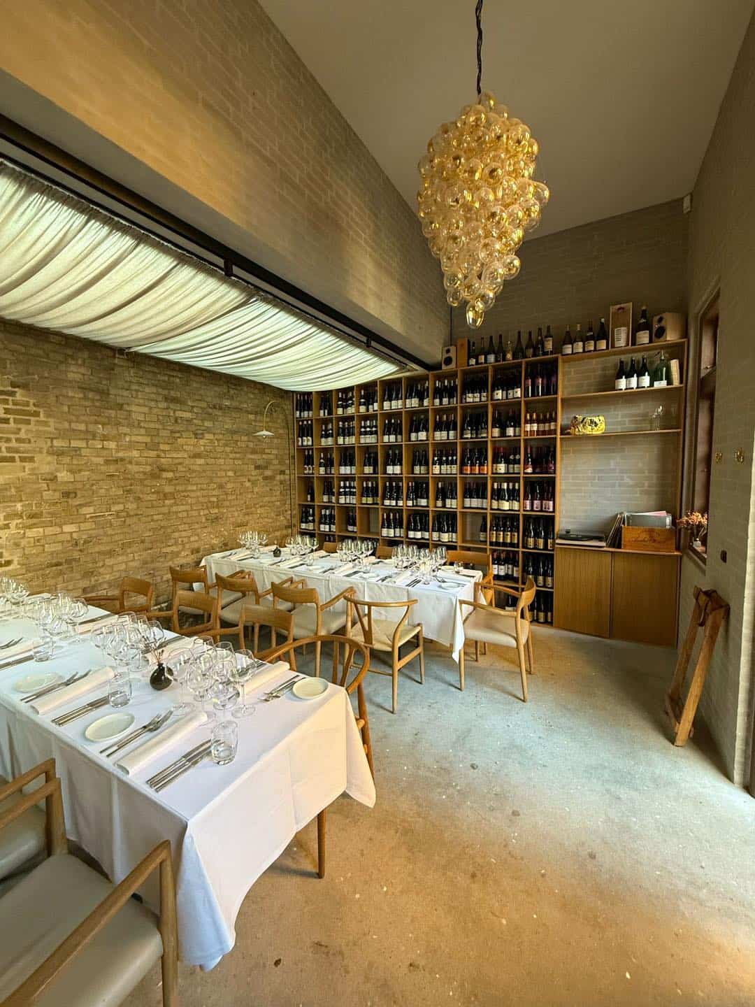 Indretning af restaurant med træborde, væg med vinreoler og elegant lysekrone.