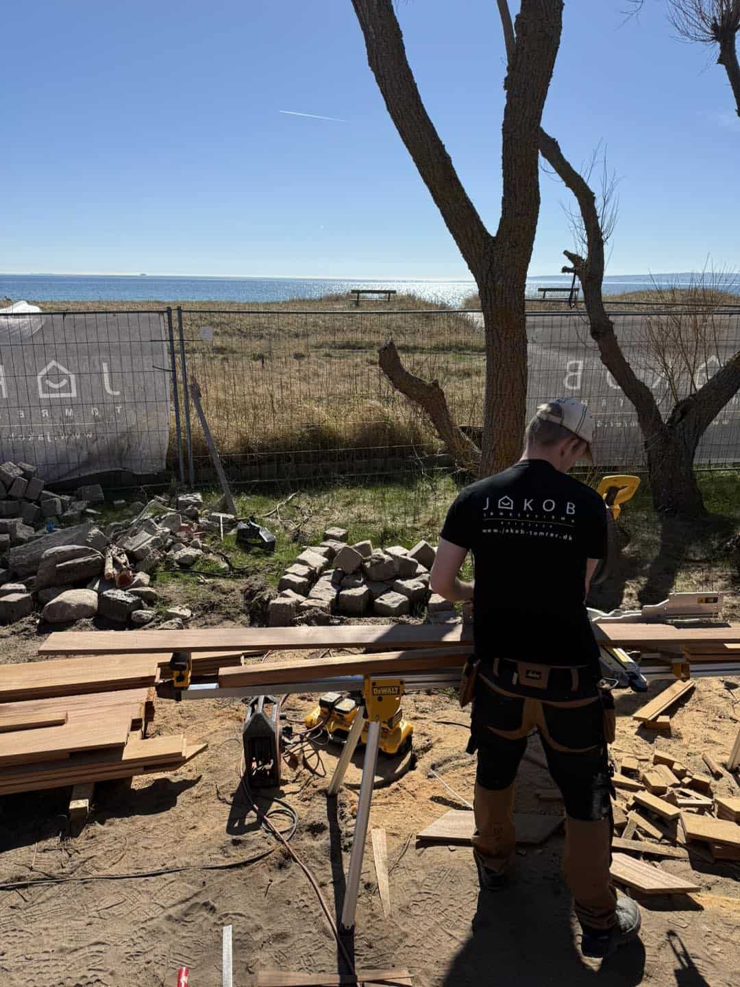 Tømrer arbejder udendørs med træmaterialer nær kysten, under blå himmel og solskin.