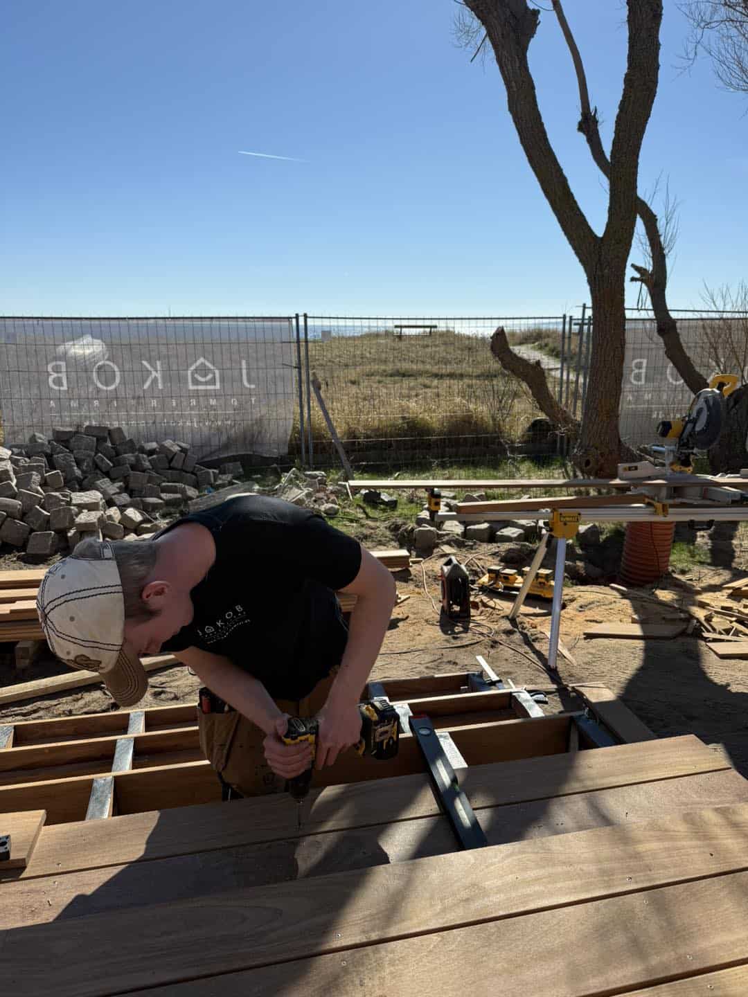 Tømrer arbejder på udendørs terrasse med træmaterialer og værktøj.