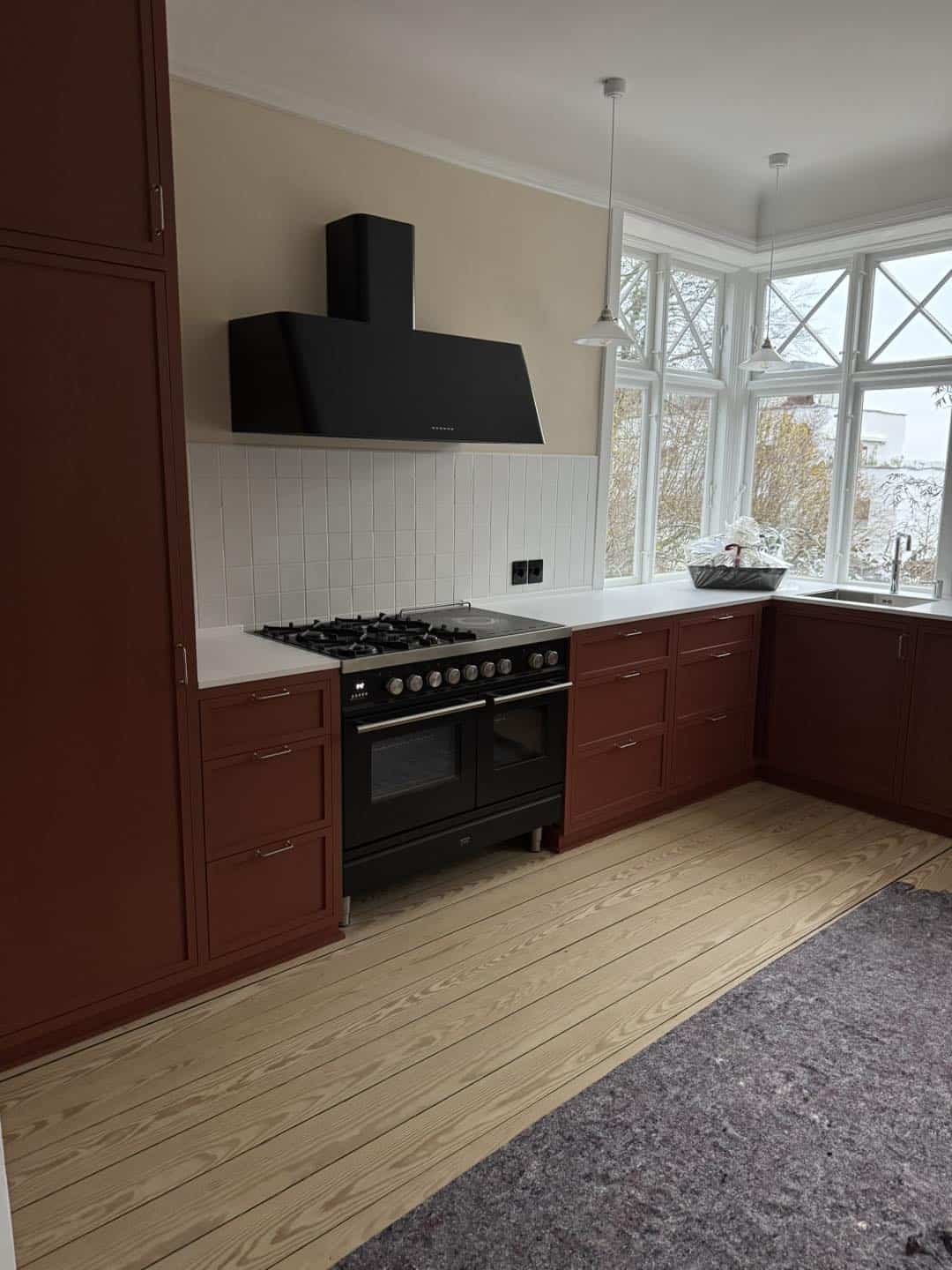 Elegant køkken med røde skabe, sort ovn og store vinduer for naturligt lys.