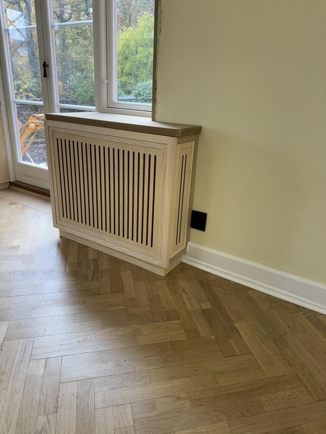 Træradiatorskjuler med lodrette lameller foran stort vindue.